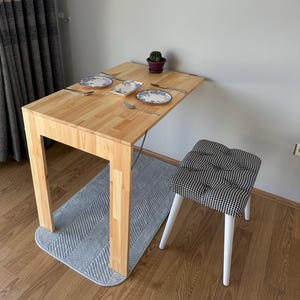 Puede incluir: Una mesa de comedor de madera clara con una parte superior rectangular y cuatro patas. Dos platos, cubiertos y un pequeño cactus en maceta están sobre la mesa. Un taburete estampado con patas blancas está cerca. Una imagen enmarcada cuelga de la pared.