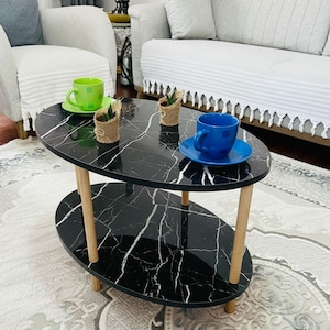 May include: Oval black marble coffee table with two tiers and light wood legs. The table is decorated with green and blue teacups and small potted plants. The table is in a living room setting with a sofa and chairs.