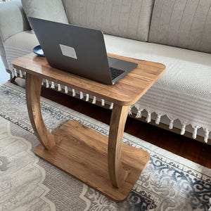 Puede incluir: Una peque&ntilde;a mesa auxiliar de madera con un port&aacute;til encima. La mesa tiene una parte superior rectangular y un estante inferior, conectados por patas curvas. La mesa es de color marr&oacute;n claro con flecos decorativos. El port&aacute;til es gris.