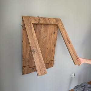 May include: A wall-mounted, folding wooden table in a natural wood grain finish. The table is partially open, with two angled support pieces extending from the main surface. The table is mounted on a gray wall.