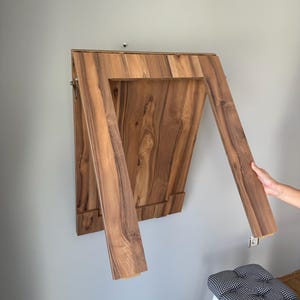 Puede incluir: Una mesa de madera plegable montada en la pared con un acabado de madera natural. La mesa es rectangular cuando está plegada y tiene dos patas de soporte con bisagras que se extienden hacia afuera. La mesa está montada en una pared gris claro.