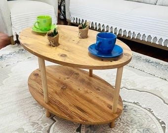 Könnte beinhalten: Ovaler Couchtisch aus Holz mit einem unteren Regal, das von vier Holzbeinen getragen wird. Der Tisch hat eine natürliche Holzoberfläche und ist mit einer grünen Tasse und Untertasse, einer blauen Tasse und Untertasse sowie kleinen Topfpflanzen verziert.
