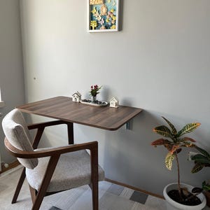 Könnte beinhalten: Ein dunkelbrauner, an der Wand montierter Klapptisch mit abgerundeter Kante, kombiniert mit einem Holzstuhl mit grau gepolstertem Sitz. Ein gerahmtes Kunstwerk mit dem Namen "ARDA" ist an der grauen Wand angebracht. Eine kleine Pflanze und dekorative Häuser stehen auf dem Tisch.
