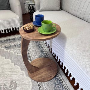 May include: A small, round wooden side table with a curved leg. The table holds a blue cup and saucer, a green cup and saucer, and a small bowl of nuts. The table is next to a light-colored sofa.