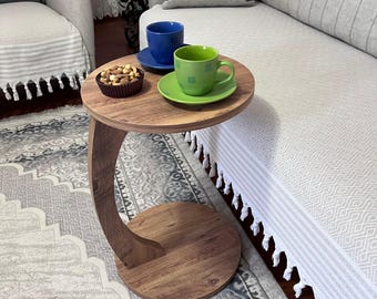 May include: A small, round wooden side table with a curved leg. The table holds a blue cup and saucer, a green cup and saucer, and a small bowl of nuts. The table is next to a light-colored sofa.