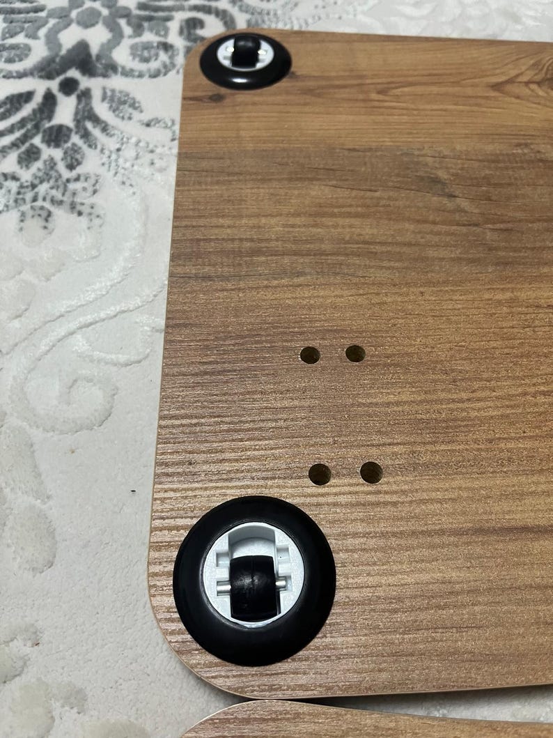 Puede incluir: Superficie de escritorio de madera con un patr&oacute;n de vetas de madera marr&oacute;n. El escritorio tiene dos ruedas negras con detalles blancos y cuatro agujeros peque&ntilde;os. El escritorio est&aacute; dise&ntilde;ado para la movilidad y la comodidad.