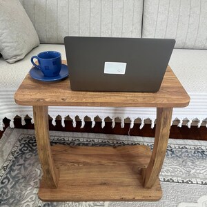 Puede incluir: Una peque&ntilde;a mesa auxiliar de madera con un port&aacute;til y una taza y un platillo azules. La mesa tiene un acabado marr&oacute;n c&aacute;lido y un estante inferior. El port&aacute;til es negro y est&aacute; abierto, y la taza y el platillo son de un azul vibrante.