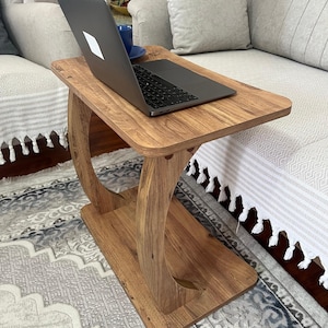 Puede incluir: Una mesa auxiliar de madera con un port&aacute;til encima. La mesa tiene una parte superior e inferior rectangulares, conectadas por soportes curvos. El port&aacute;til est&aacute; abierto y hay una taza azul cerca. La mesa es de madera de color marr&oacute;n claro.