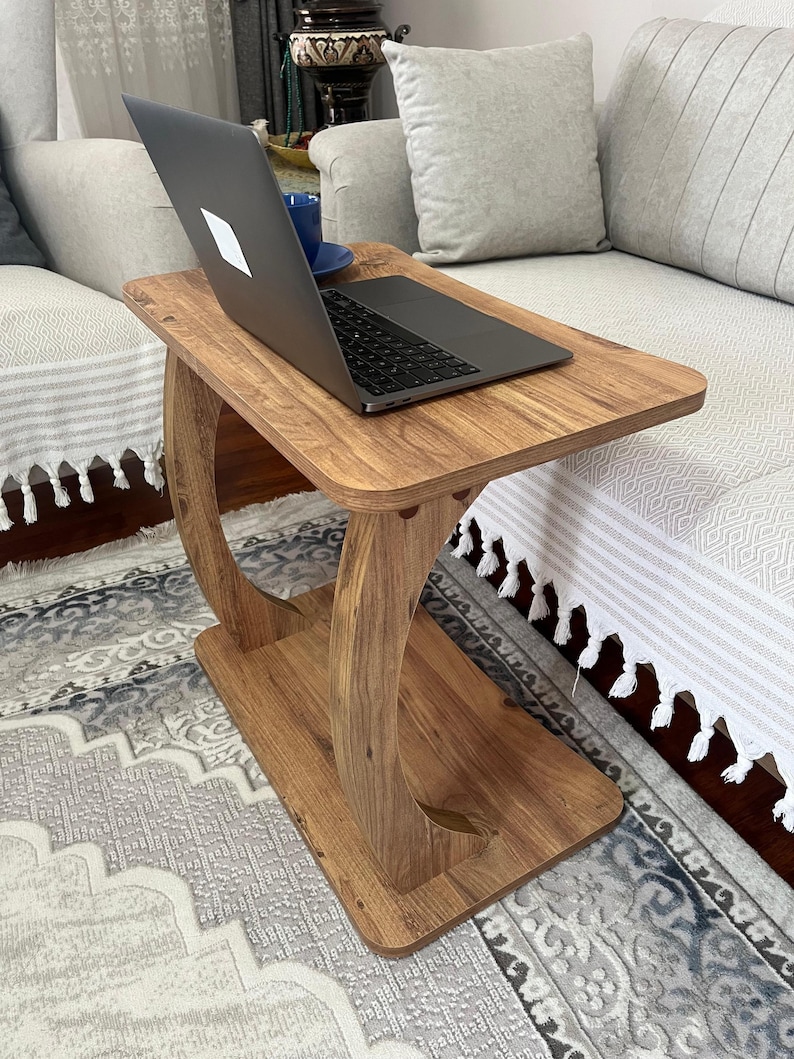 Puede incluir: Una mesa auxiliar de madera con un port&aacute;til encima. La mesa tiene una parte superior e inferior rectangulares, conectadas por soportes curvos. El port&aacute;til est&aacute; abierto y hay una taza azul cerca. La mesa es de madera de color marr&oacute;n claro.