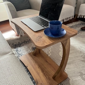 Puede incluir: Una mesa de madera para port&aacute;til con un dise&ntilde;o curvo. Una computadora port&aacute;til y una taza y platillo azules est&aacute;n sobre la mesa. La mesa tiene un acabado de vetas de madera marr&oacute;n claro y est&aacute; sostenida por una pata curva.