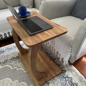 Puede incluir: Una mesa auxiliar de madera con un port&aacute;til y una taza de t&eacute; azul y un platillo. La mesa tiene un acabado marr&oacute;n claro y un dise&ntilde;o de pata curvada &uacute;nico. El port&aacute;til est&aacute; abierto, mostrando una pantalla oscura.