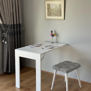 Könnte beinhalten: Ein weißer, an der Wand befestigter Klapptisch mit einem weißen Bein. Ein kleiner Hocker mit schwarz-weiß gemustertem Sitz und weißen Beinen steht daneben. Zwei Teller mit Besteck sind auf dem Tisch. Ein gerahmtes Bild und eine Vase sind an der Wand.