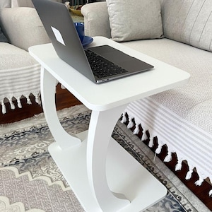 Puede incluir: Una mesa auxiliar blanca con una parte superior rectangular y soportes curvos. Una computadora port&aacute;til y una taza azul est&aacute;n sobre la mesa. La mesa est&aacute; al lado de un sof&aacute; con cojines grises y una alfombra estampada.