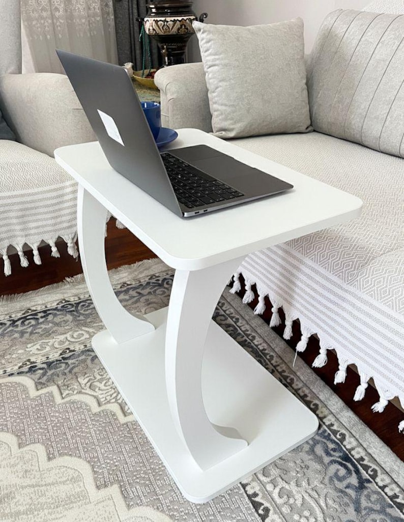 Puede incluir: Una mesa auxiliar blanca con una parte superior rectangular y soportes curvos. Una computadora port&aacute;til y una taza azul est&aacute;n sobre la mesa. La mesa est&aacute; al lado de un sof&aacute; con cojines grises y una alfombra estampada.