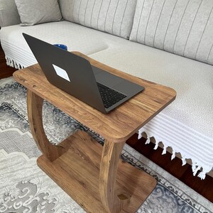 Puede incluir: Una mesa auxiliar de madera con un port&aacute;til encima. La mesa tiene una parte superior rectangular y un estante inferior, conectados por soportes curvos. El port&aacute;til es negro y se ve una taza azul. La mesa est&aacute; sobre una alfombra estampada.