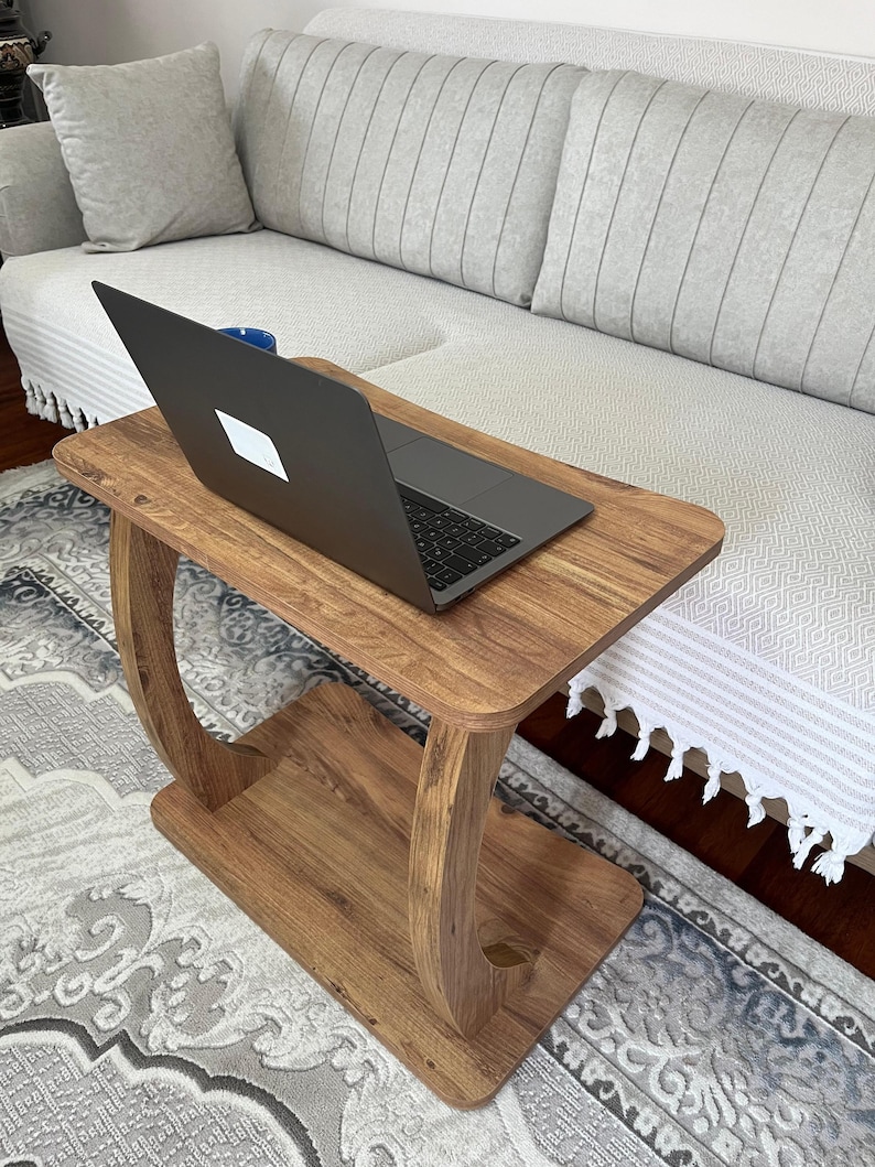 Puede incluir: Una mesa auxiliar de madera con un port&aacute;til encima. La mesa tiene una parte superior rectangular y un estante inferior, conectados por soportes curvos. El port&aacute;til es negro y se ve una taza azul. La mesa est&aacute; sobre una alfombra estampada.