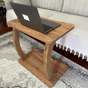 Puede incluir: Una mesa de madera para port&aacute;til con acabado marr&oacute;n c&aacute;lido. La mesa tiene una parte superior rectangular y un estante inferior, sostenidos por patas curvas. Un ordenador port&aacute;til negro est&aacute; colocado sobre la mesa. La mesa es pr&aacute;ctica y funcional.