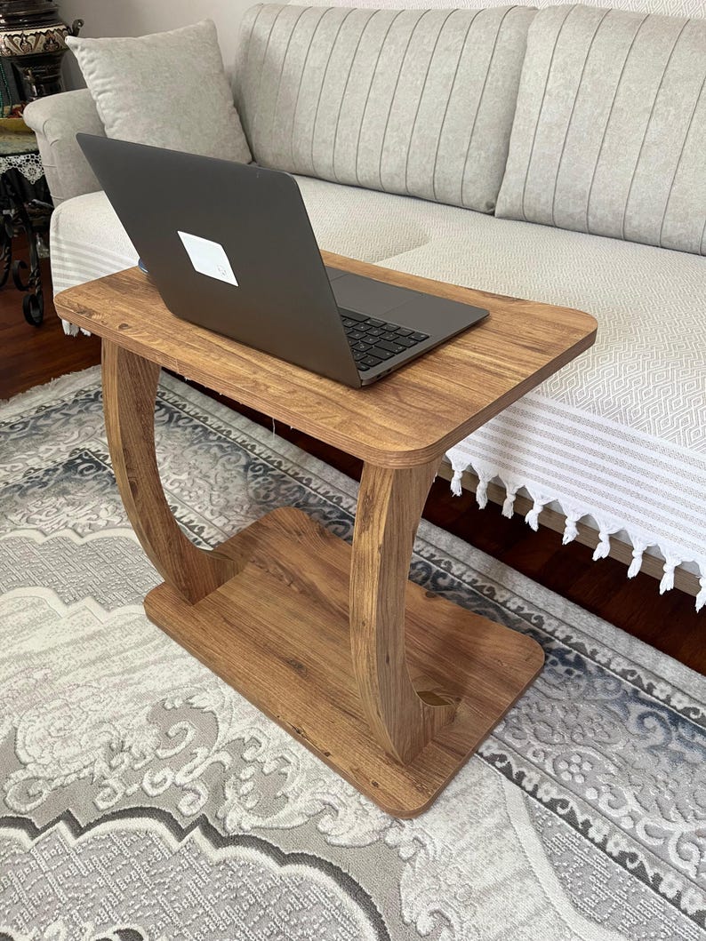 Puede incluir: Una mesa de madera para port&aacute;til con acabado marr&oacute;n c&aacute;lido. La mesa tiene una parte superior rectangular y un estante inferior, sostenidos por patas curvas. Un ordenador port&aacute;til negro est&aacute; colocado sobre la mesa. La mesa es pr&aacute;ctica y funcional.