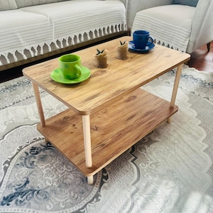 May include: A two-tiered wooden coffee table with a light brown finish. The table has a rectangular top and a lower shelf, supported by four cylindrical legs. Two small potted plants and two cups and saucers are on the top surface.
