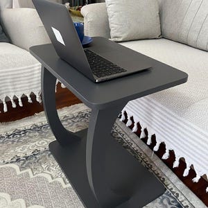 Puede incluir: Una mesa auxiliar gris oscuro con base curva, que sostiene un port&aacute;til y una taza azul. La mesa est&aacute; colocada junto a un sof&aacute; gris claro con cojines decorativos. El port&aacute;til est&aacute; abierto, mostrando un logotipo blanco.