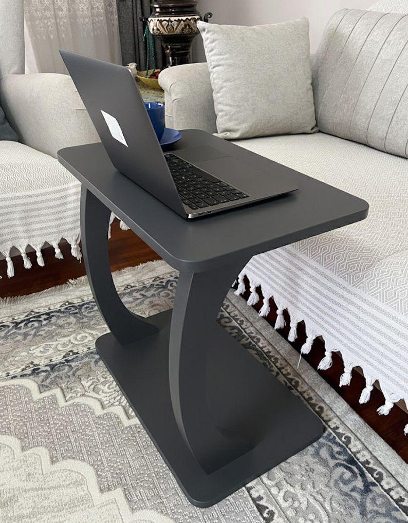 Puede incluir: Una mesa auxiliar gris oscuro con base curva, que sostiene un port&aacute;til y una taza azul. La mesa est&aacute; colocada junto a un sof&aacute; gris claro con cojines decorativos. El port&aacute;til est&aacute; abierto, mostrando un logotipo blanco.