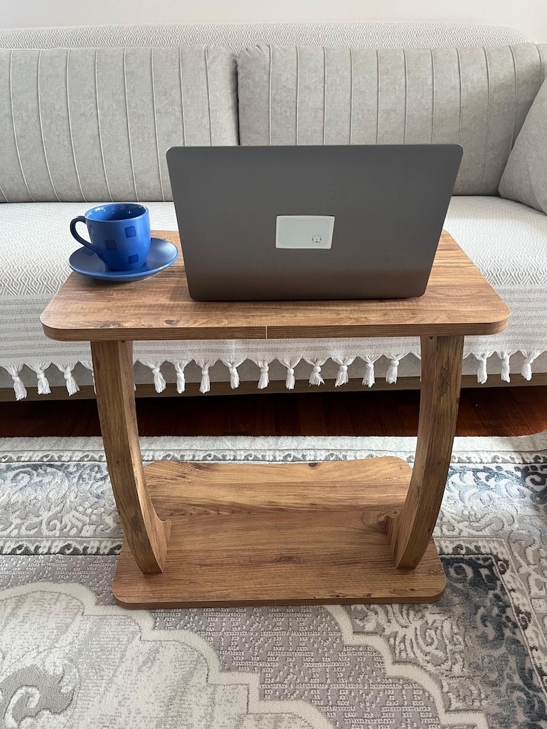 Puede incluir: Una peque&ntilde;a mesa auxiliar de madera con un port&aacute;til y una taza y un platillo azules. La mesa tiene un estante inferior y patas curvas. El port&aacute;til est&aacute; cerrado y tiene una pegatina blanca en la parte trasera.