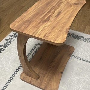 Puede incluir: Una mesa auxiliar de madera con un soporte curvo y dos superficies rectangulares. La mesa tiene un acabado de veta de madera marr&oacute;n claro. La mesa mide aproximadamente 61 cm de largo, 30 cm de ancho y 51 cm de alto.