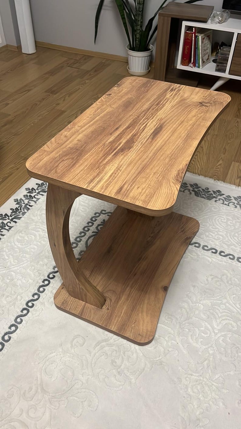 Puede incluir: Una mesa auxiliar de madera con un soporte curvo y dos superficies rectangulares. La mesa tiene un acabado de veta de madera marr&oacute;n claro. La mesa mide aproximadamente 61 cm de largo, 30 cm de ancho y 51 cm de alto.