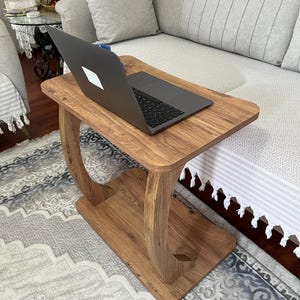 May include: A wooden side table with a laptop on top. The table has a rectangular top and a lower shelf, connected by curved supports. The wood has a warm, natural finish. The laptop is closed and has a white logo.