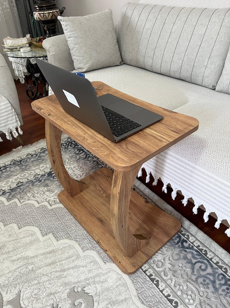 Puede incluir: Una mesa auxiliar de madera con un port&aacute;til encima. La mesa tiene una parte superior rectangular y un estante inferior, conectados por soportes curvos. La madera tiene un acabado c&aacute;lido y natural. El port&aacute;til est&aacute; cerrado.