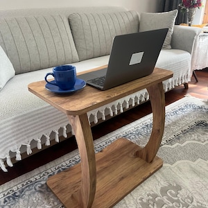 Puede incluir: Una mesa auxiliar de madera con un dise&ntilde;o de patas curvas. Una taza y un platillo azules est&aacute;n sobre la mesa, junto con un ordenador port&aacute;til. La mesa es de madera de color marr&oacute;n claro y est&aacute; colocada frente a un sof&aacute; claro.