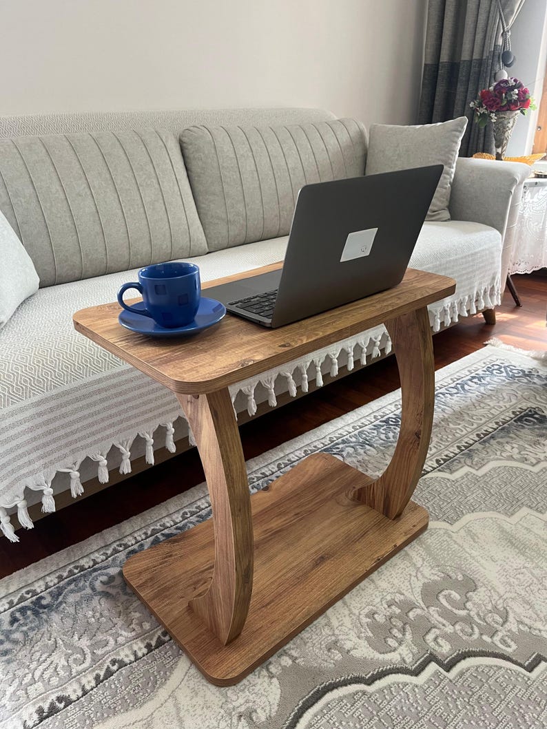 Puede incluir: Una mesa auxiliar de madera con un dise&ntilde;o de patas curvas. Una taza y un platillo azules est&aacute;n sobre la mesa, junto con un ordenador port&aacute;til. La mesa es de madera de color marr&oacute;n claro y est&aacute; colocada frente a un sof&aacute; claro.
