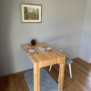 May include: A light-colored wooden dining table with four legs, set with two blue and white patterned plates, a small rectangular dish, and silverware. A small potted cactus sits on the table, and a framed print hangs on the wall above.