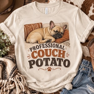 Könnte beinhalten: Ein cremefarbenes Kurzarm-T-Shirt mit einer Grafik einer falbfarbenen Französischen Bulldogge, die auf einem braunen Kissen schläft. Unter dem Hund steht der Text "PROFESSIONAL COUCH POTATO" in braunen und schwarzen Buchstaben mit Pfotenabdruck.