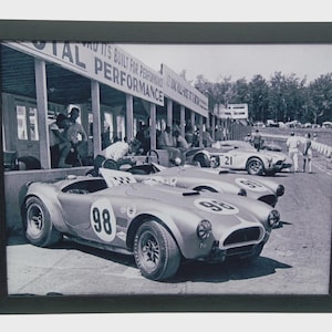 May include: Framed black and white photograph of vintage race cars in a pit area. The cars are light-colored with racing numbers. The photo includes a pit area with people and the words "TOTAL PERFORMANCE" visible.