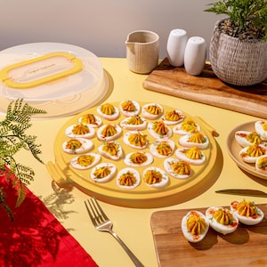 Op de afbeelding: Een gele serveerschaal gevuld met gevulde eieren, gegarneerd met kruiden en paprika. Een doorzichtige plastic container met een gele handgreep staat op de achtergrond. Andere serveerschalen en een kleine plant zijn ook zichtbaar.