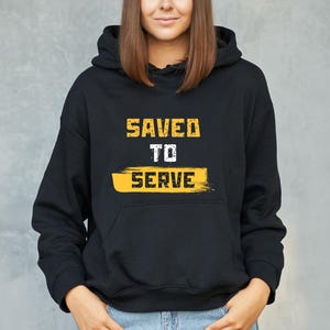 Könnte beinhalten: Schwarzer Kapuzenpullover mit dem Text "SAVED TO SERVE" in Gelb und Weiß. Der Text befindet sich über einem gelben horizontalen Rechteck. Der Kapuzenpullover hat eine Kapuze mit Kordelzug und eine Vordertasche.