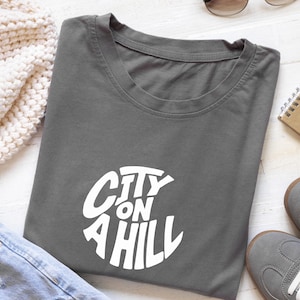 May include: A gray t-shirt with the white text "CITY ON A HILL" in a circular design. The shirt is laid flat with a pair of gray sneakers, a beige notebook, and a cream-colored knit sweater nearby.