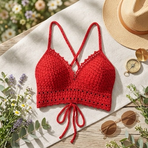 Può includere: Un top all'uncinetto rosso acceso con schiena incrociata e chiusura con lacci. Il top presenta un motivo a punto strutturato e bordi delicati. Nell'immagine sono visibili anche un cappello di paglia, occhiali da sole e una bussola.