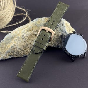 May include: Olive green leather watch strap with a rose gold buckle. The strap is stitched with a matching thread. A black smartwatch with a reflective screen is next to the strap. A stone and a ball of twine are in the background.