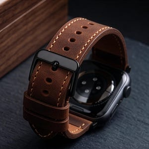Könnte beinhalten: Ein braunes Lederarmband mit sichtbaren hellen Nähten und einer schwarzen Metallschnalle. Das Armband ist an einem schwarzen Smartwatch-Gehäuse befestigt und zeigt die Sensoranordnung auf der Rückseite sowie eine matte Oberfläche.
