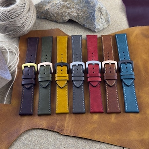May include: A collection of seven leather watch straps in various colors, including dark purple, olive green, yellow, gray, red, brown, and teal. Each strap features a contrasting stitch and a metal buckle in gold, silver, or black.