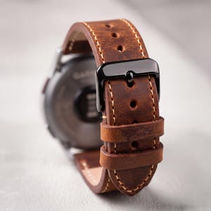 Puede incluir: Una correa de reloj de cuero marrón con costuras claras visibles y una hebilla de metal negro. La correa presenta múltiples orificios de ajuste y dos trabillas de cuero, unida a la parte posterior de una caja de reloj circular oscura.