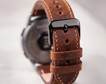 Può includere: Un cinturino per orologio in pelle marrone con cuciture chiare visibili e una fibbia in metallo nero. Il cinturino presenta diversi fori di regolazione e due passanti in pelle, fissato al retro di una cassa dell'orologio circolare scura.