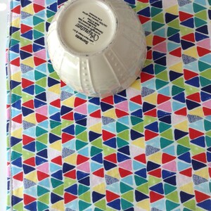 May include: A white ceramic bowl with a geometric pattern on the bottom. The bowl is sitting on a colorful fabric placemat with a pattern of small triangles in shades of red, blue, green, yellow, and pink.