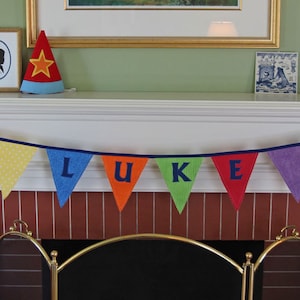 May include: A white fireplace mantel with a banner that reads "LUKE" in colorful letters. The banner is made of fabric and is strung with colorful yarn. There is a red and white star-shaped hat on the mantel, as well as a framed picture and a small wooden horse figurine.