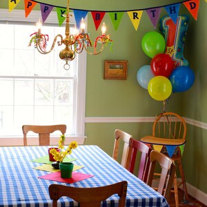 May include: A green dining room with a blue and white checkered tablecloth on the table. There are four chairs around the table, and a small wooden chair with a banner that says "Happy Birthday" hanging from it. There are colorful balloons in the background.