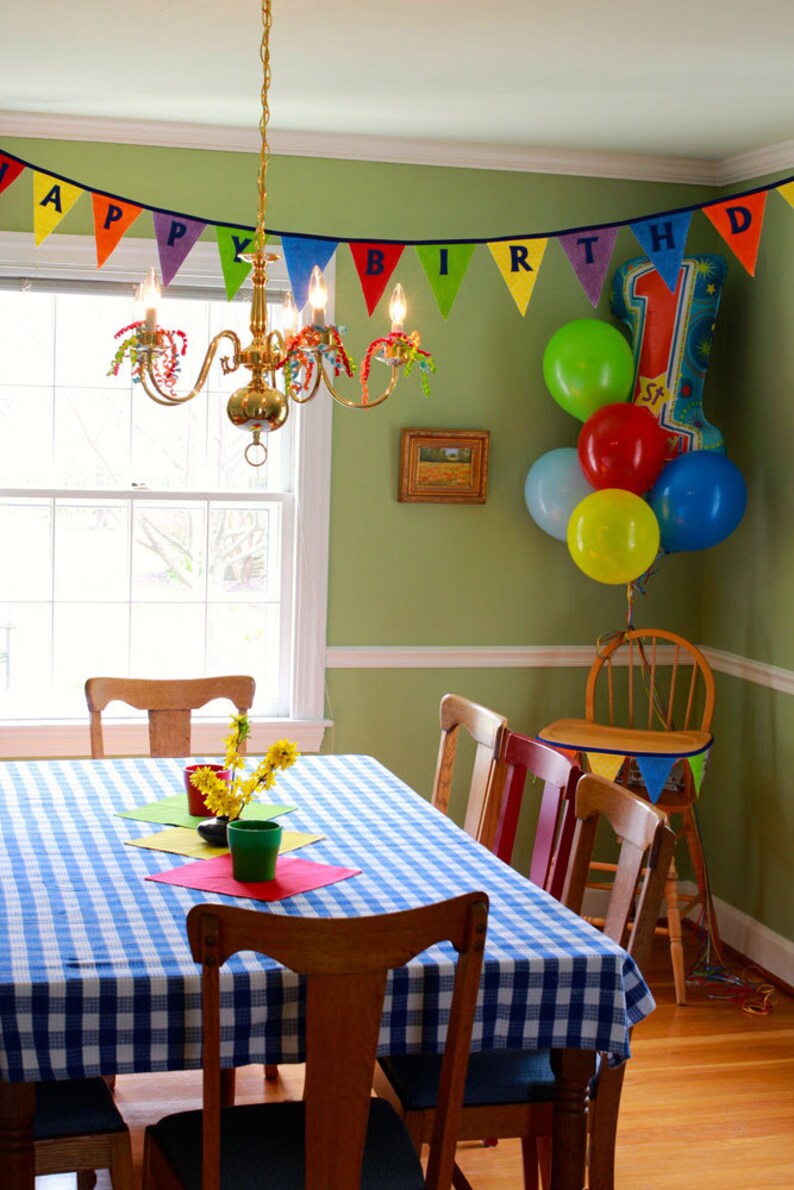 May include: A green dining room with a blue and white checkered tablecloth on the table. There are four chairs around the table, and a small wooden chair with a banner that says "Happy Birthday" hanging from it. There are colorful balloons in the background.