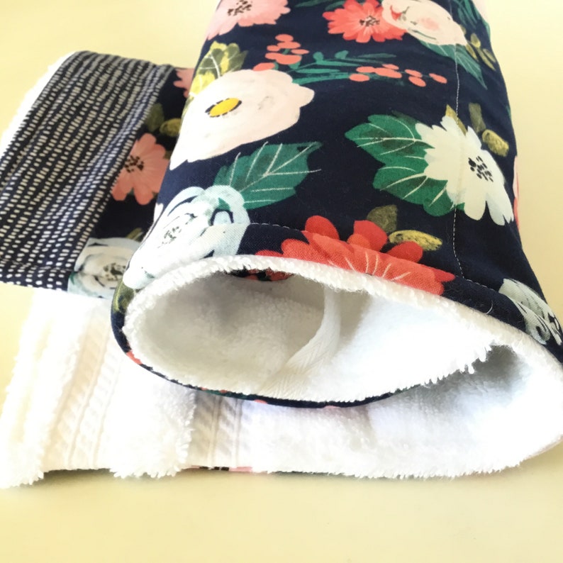 May include: A set of handmade oven mitts and a towel. The oven mitts feature a navy blue floral pattern with pink and white flowers and green leaves. The interior is white. The towel is white with a textured pattern and a black and white patterned border.