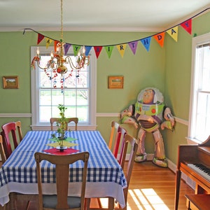 May include: A green and white checkered tablecloth covers a table set for a birthday party. A colorful banner with the words "Happy Birthday" hangs above the table. A large inflatable Buzz Lightyear toy stands in the corner of the room.